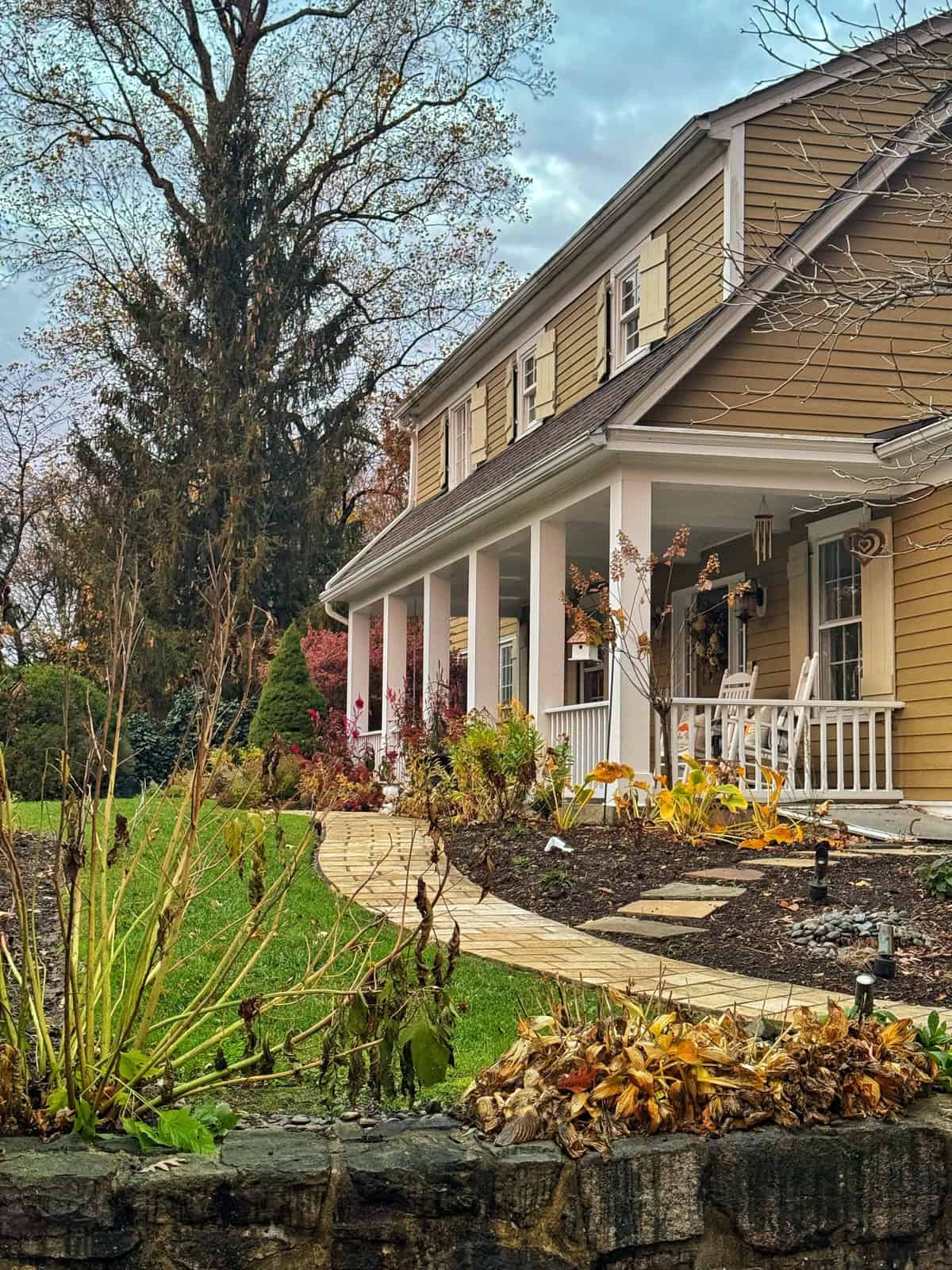 A tan two-story house with white trim and a large front porch, featuring rocking chairs and potted plants. A curved stone path leads through a garden with autumn plants, and tall trees stand in the background.