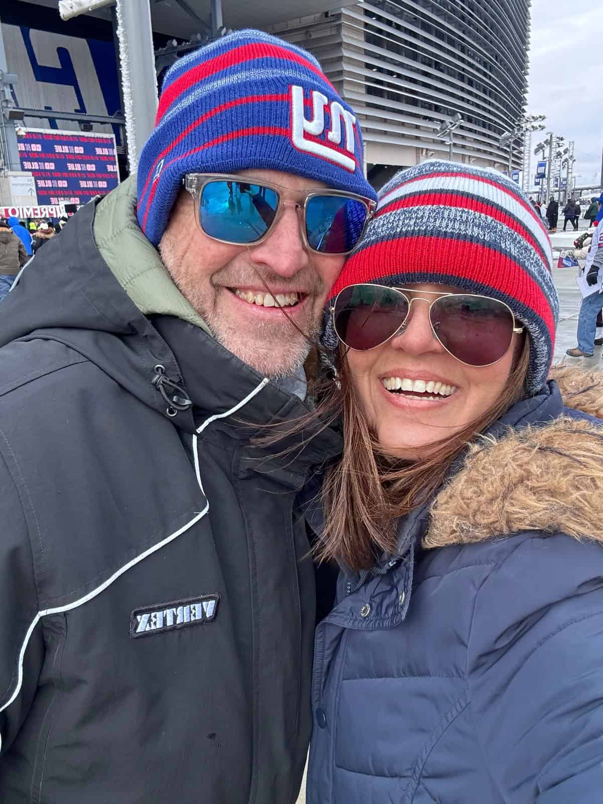 A smiling couple wearing winter jackets, knitted hats, and sunglasses pose for a selfie outside a stadium. The man&rsquo;s hat has a New York Giants logo, and both appear happy and bundled up for cold weather.