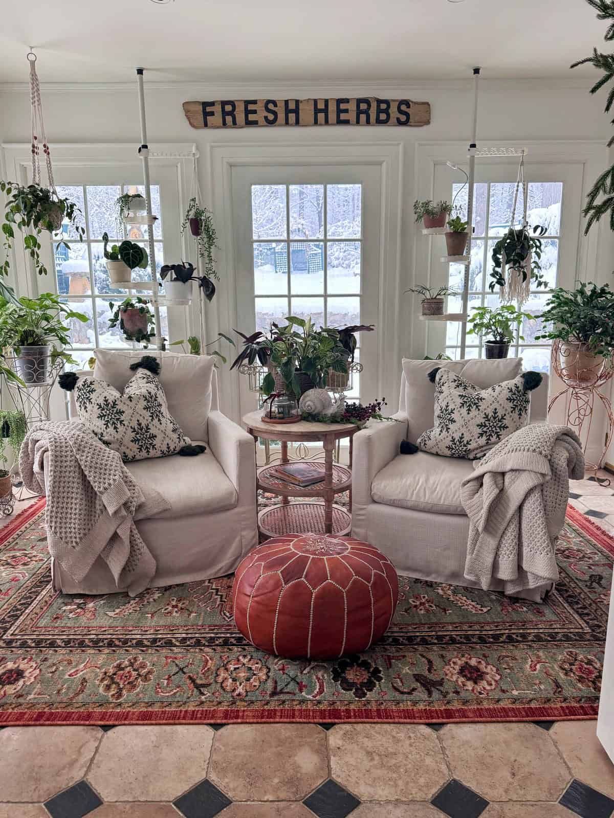Two white armchairs with knit blankets and patterned pillows sit on a colorful rug, surrounded by houseplants. A round brown ottoman is in front, and a sign above the glass doors reads "FRESH HERBS.