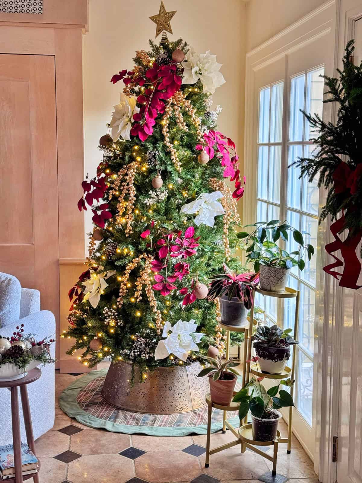A decorated Christmas tree with red and white poinsettia flowers, gold ornaments, and lights stands by a window. Several potted plants are arranged on a gold stand and floor nearby, with sunlight streaming in.