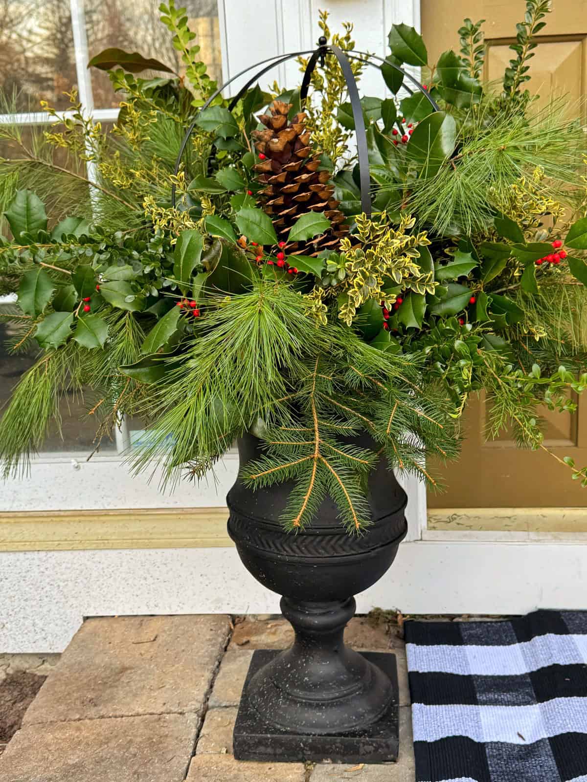 A black urn planter filled with evergreen branches, holly leaves, red berries, yellow sprigs, and a large pinecone sits on a doorstep beside a gold door and a black-and-white striped doormat.