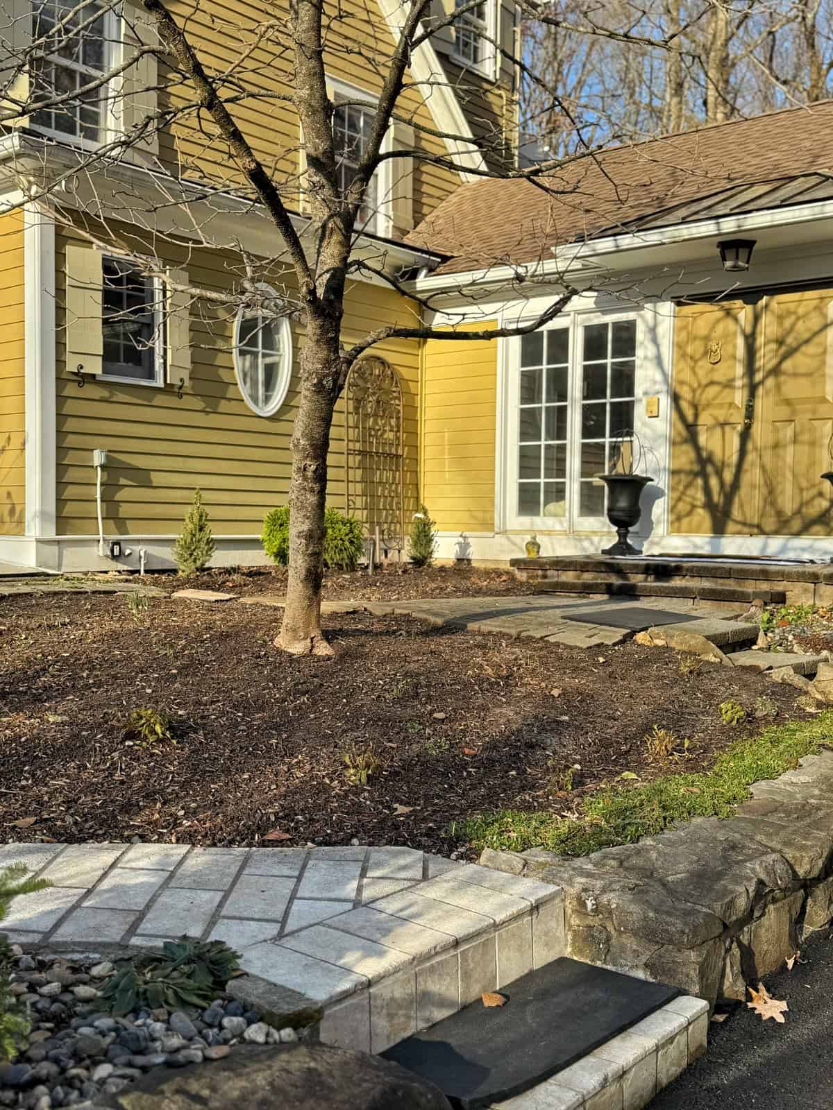 A yellow house with white trim, a bare tree in the front yard, a stone path, small plants, and a black urn near a set of white French doors. Sunlight casts shadows across the scene.