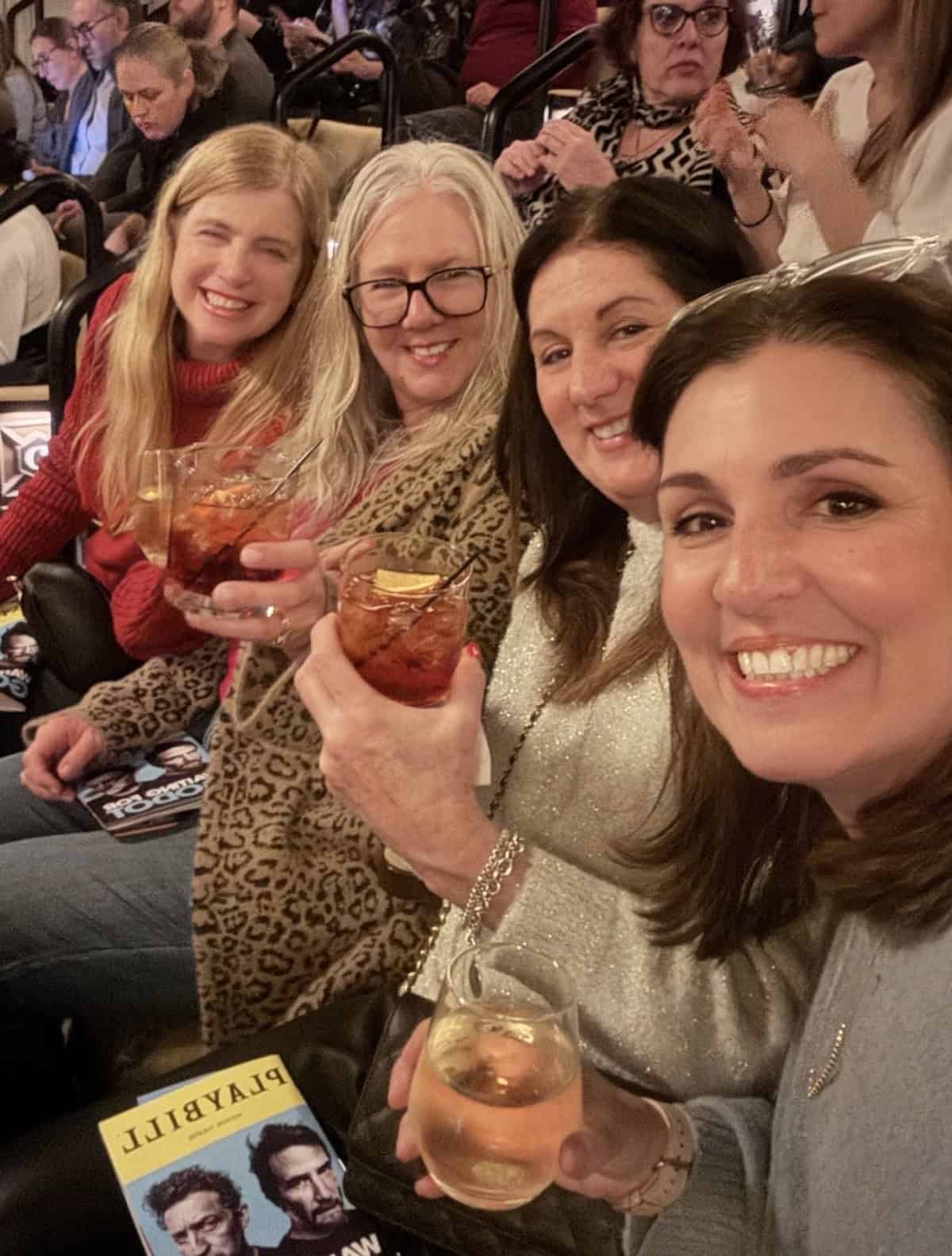 Four women sitting in theater seats, smiling and holding drinks. Playbills for &ldquo;Sweeney Todd&rdquo; are visible on their laps. Other theatergoers are seated in the background.