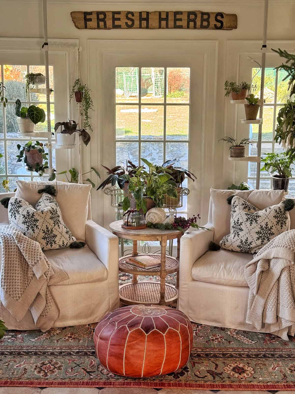 Two cozy armchairs with patterned pillows and blankets sit by a window, surrounded by potted plants. A round ottoman and a small table with plants are in front, and a “Fresh Herbs” sign hangs above the window.