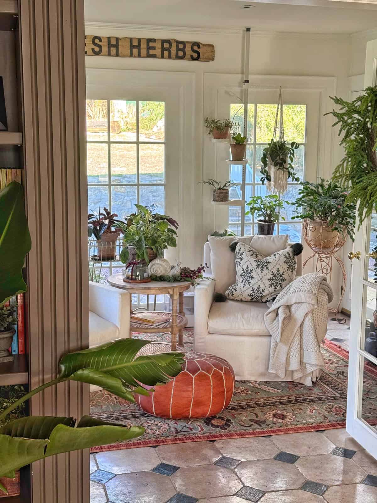 A cozy sunroom with large windows, filled with potted and hanging plants, a white armchair with a floral pillow and blanket, a round ottoman, a patterned rug, and a wooden "FRESH HERBS" sign on the wall.