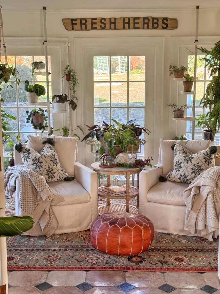 Two cozy white armchairs with patterned pillows and blankets sit by large windows, surrounded by potted plants and greenery. A round leather pouf and a small table with plants are between the chairs. A "Fresh Herbs" sign hangs above.