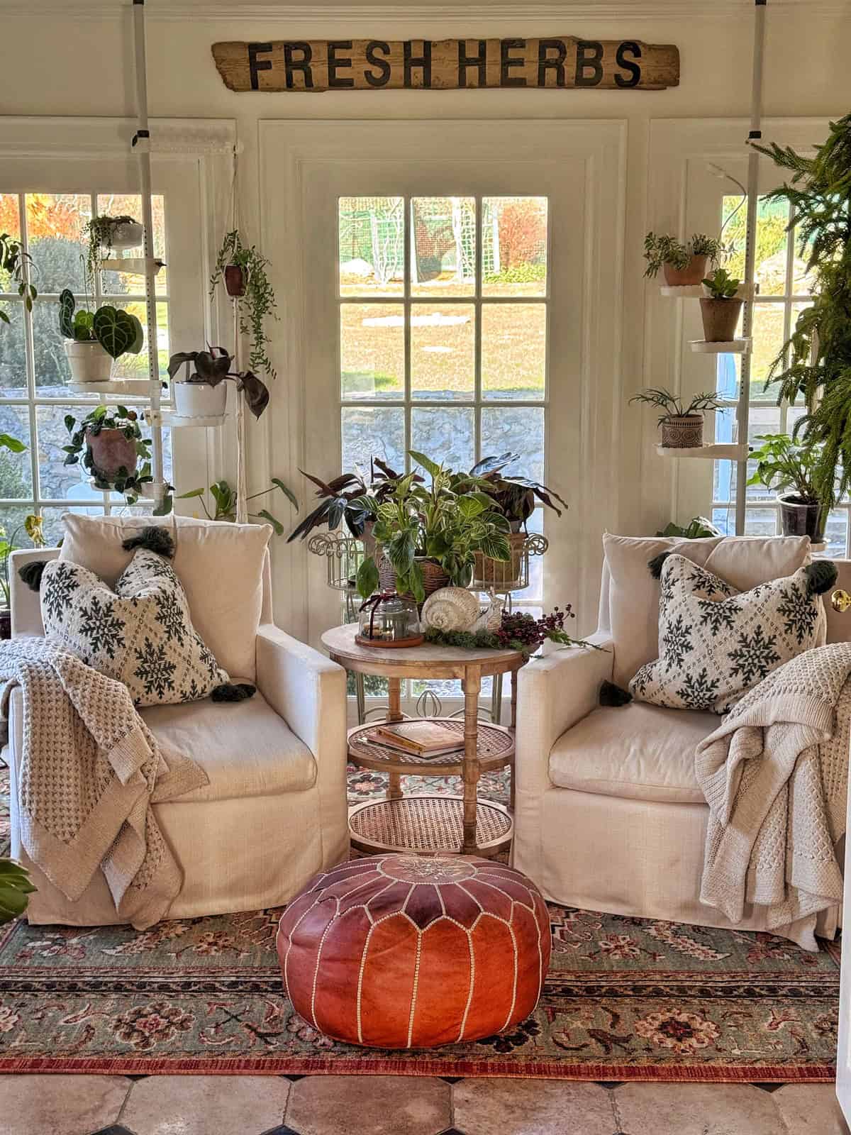 Two cozy armchairs with patterned pillows and throws sit by a window, surrounded by potted plants. A round ottoman and small side table add warmth to the space. A wooden &ldquo;Fresh Herbs&rdquo; sign hangs above the window.
