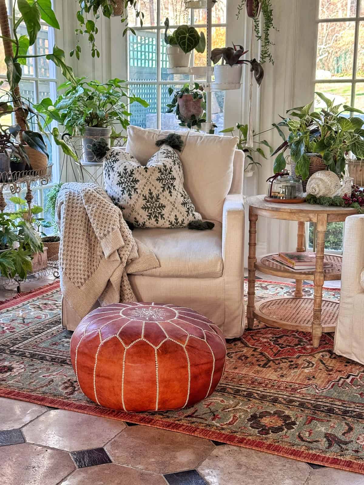 A cozy sunlit room with a cream armchair, snowflake-patterned pillows, a knit blanket, a round brown leather pouf, potted plants, a teapot on a small table, and layered patterned rugs on the floor.