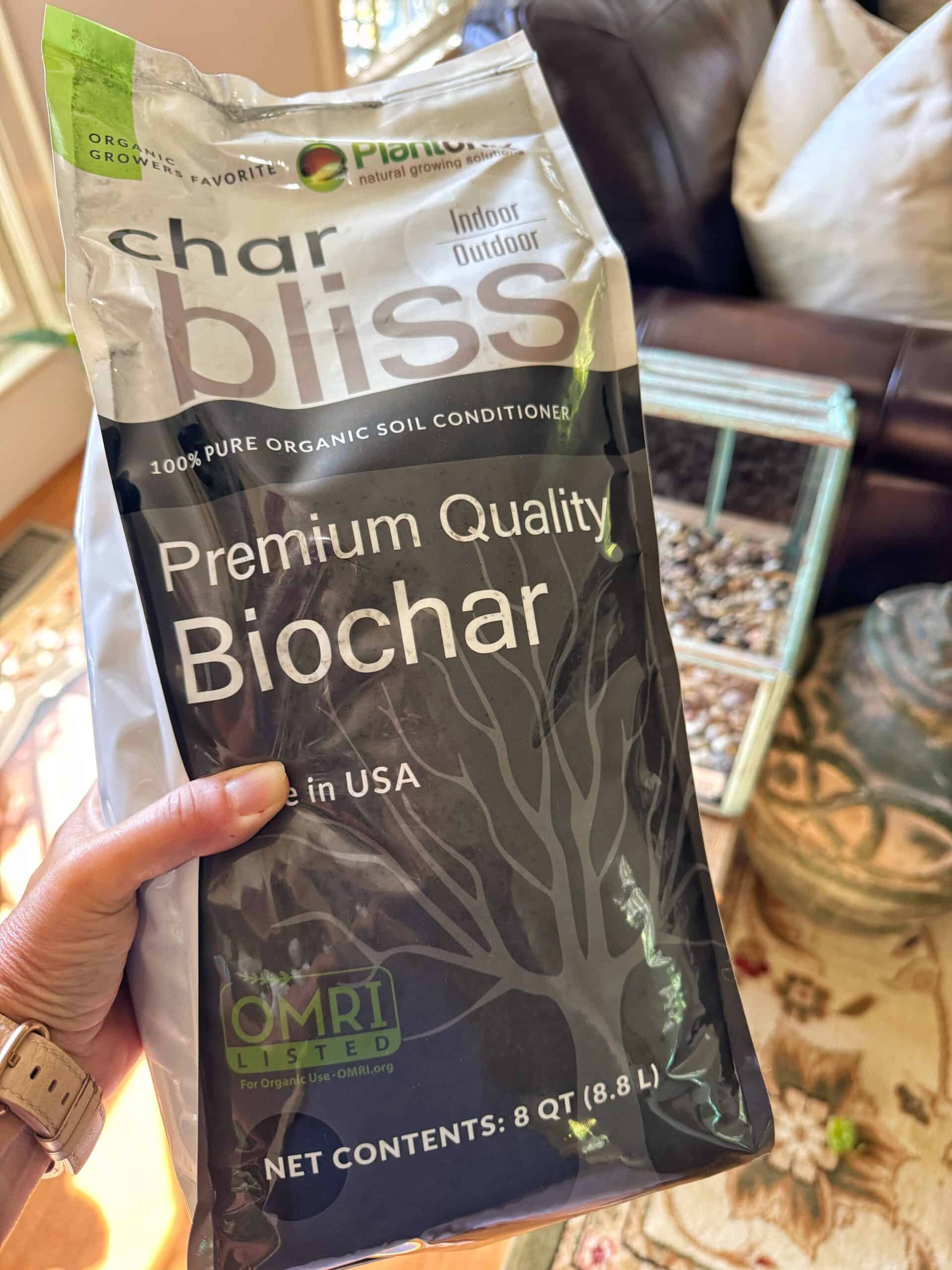 A hand holding a bag of char bliss Premium Quality Biochar soil conditioner, labeled as 100% pure organic and OMRI listed, with indoor and outdoor use indicated. A glass container with pebbles is in the background.