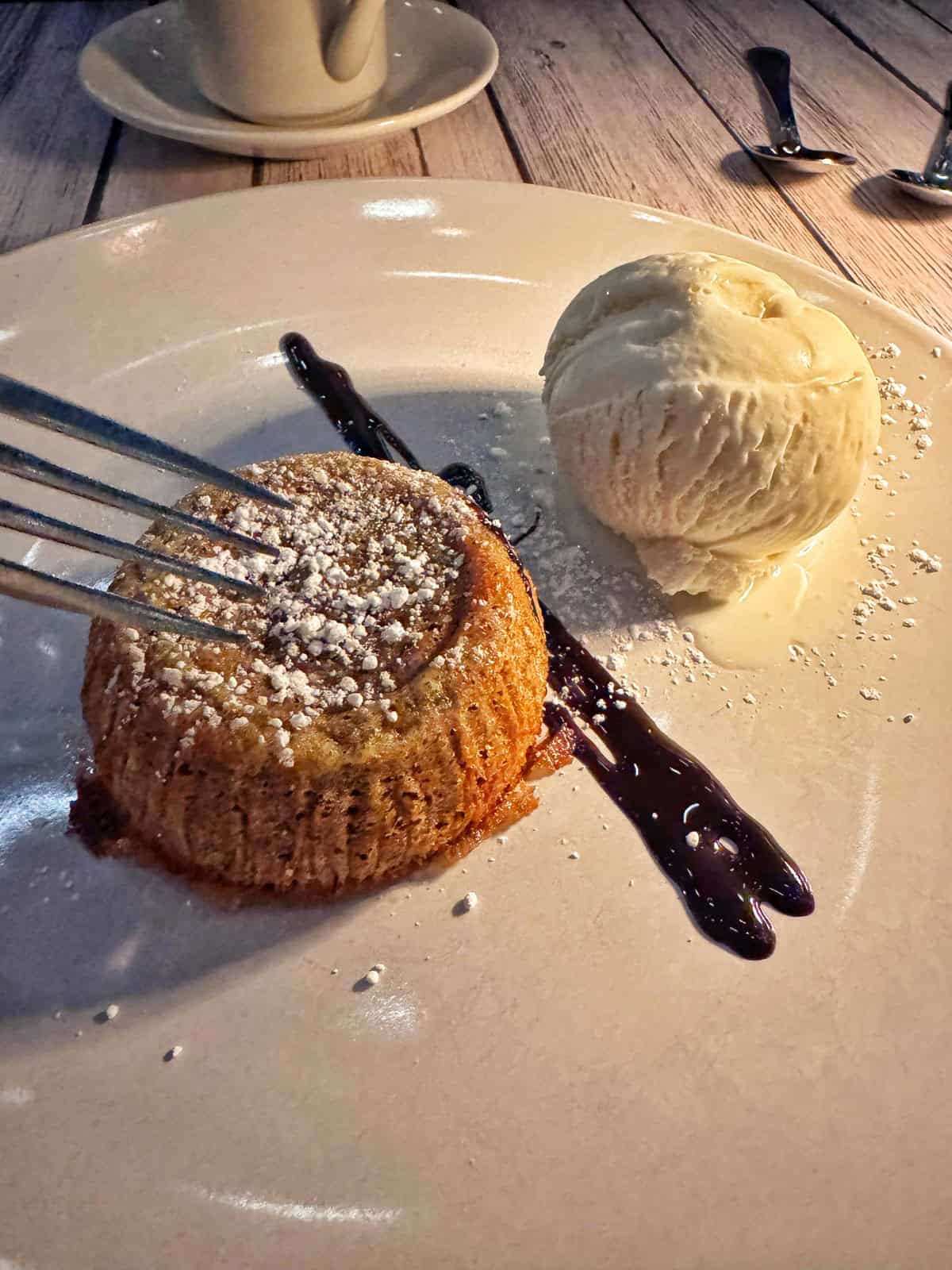 A dessert plate with a small cake topped with powdered sugar and a fork pressing on it, next to a scoop of vanilla ice cream and a streak of chocolate sauce, on a wooden table.