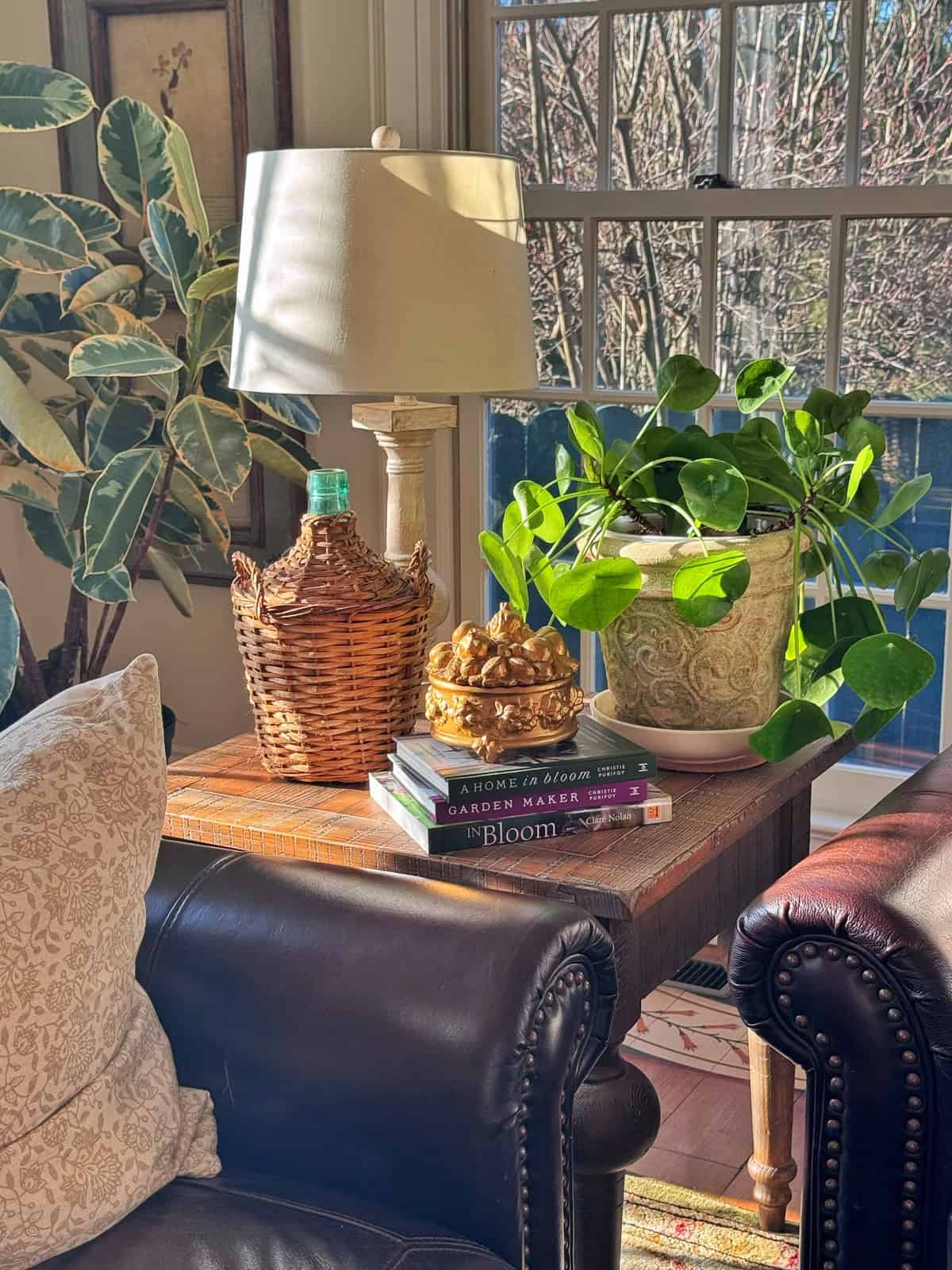 A sunlit side table by a window holds a wicker lamp, a plant in a decorative pot, a wicker-covered bottle, a small golden ornament, and stacked gardening books between two leather chairs.