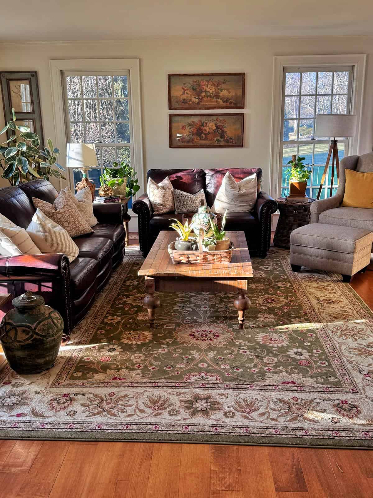 A sunlit living room with two leather sofas, a gray armchair, a wooden coffee table with plants, decorative pillows, a patterned rug, large windows, and framed art on the wall. Plants sit by the windows.
