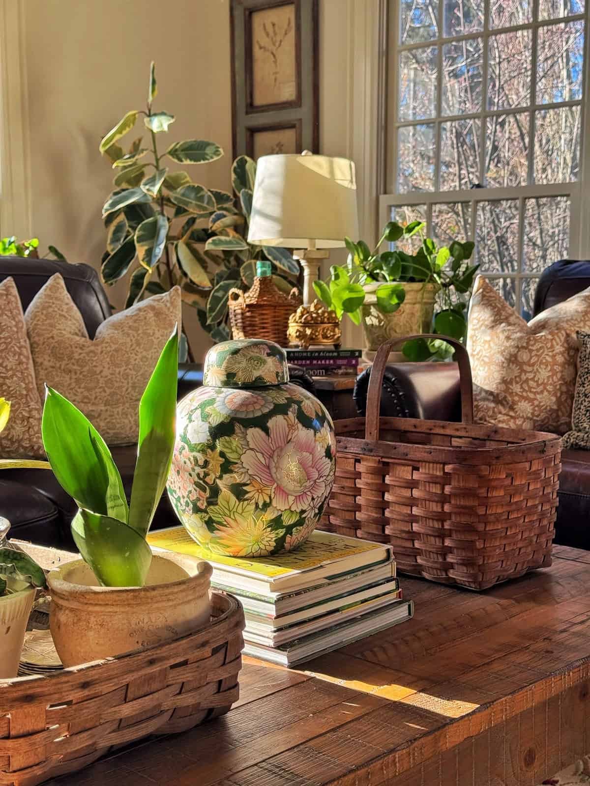 A sunlit living room with a wooden coffee table holding a floral vase, a potted plant, two woven baskets, and stacked books. Behind, a couch with patterned pillows and large windows with leafy plants.