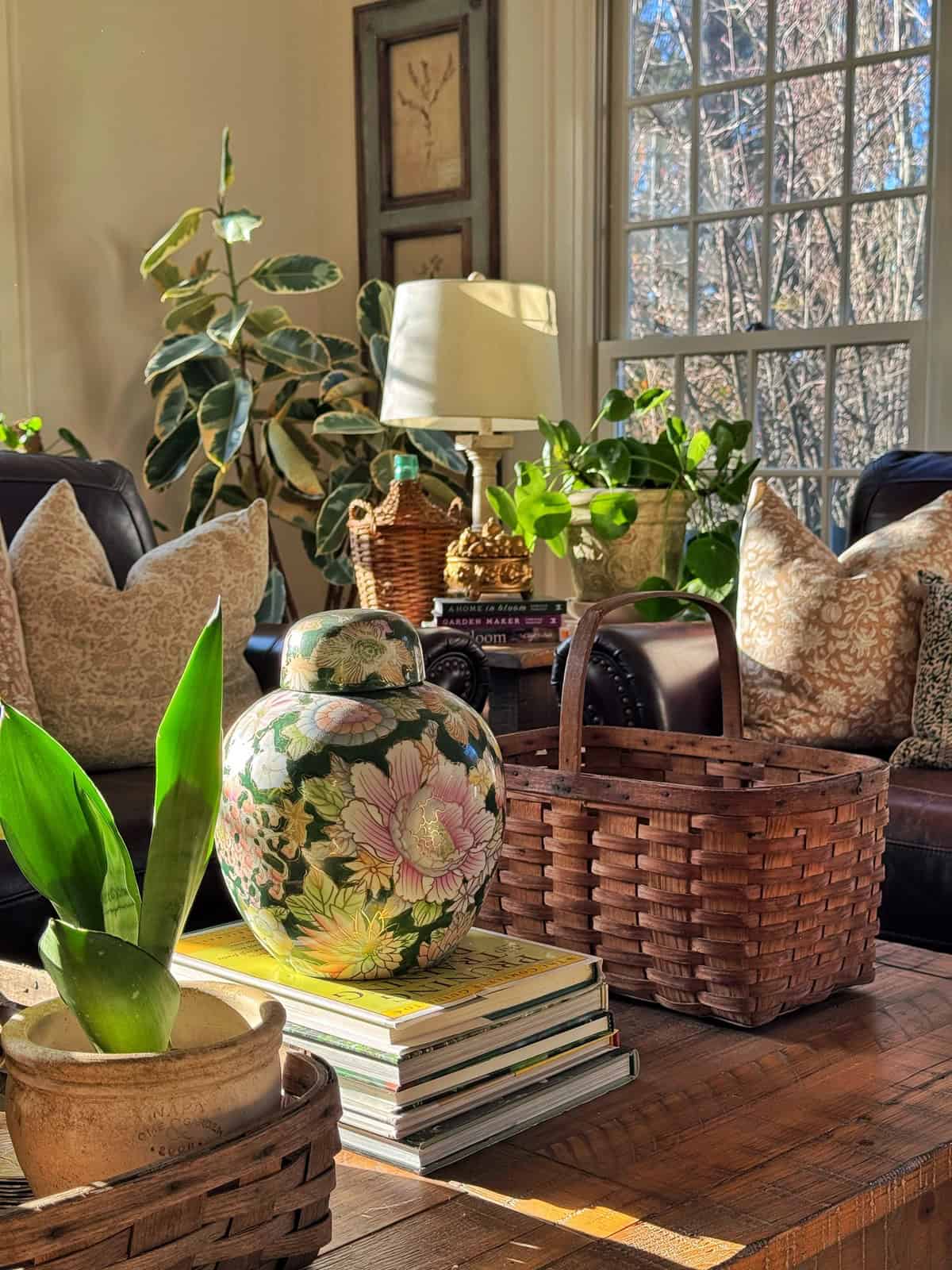 A sunlit living room with green plants, patterned cushions on a dark sofa, a floral ceramic vase, stacked books, baskets, and a large window allowing natural light to fill the cozy, inviting space.