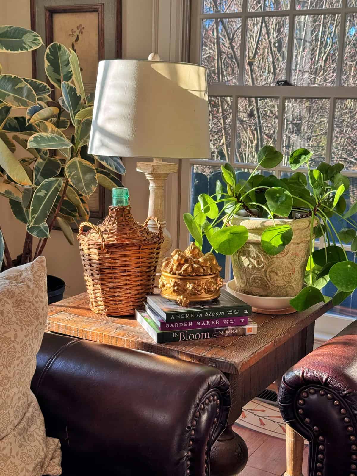A sunlit wooden side table holds a white lamp, a wicker-covered bottle, a gold decorative crown, two potted plants, and stacked garden books, set next to a leather chair and large window with leafy trees outside.