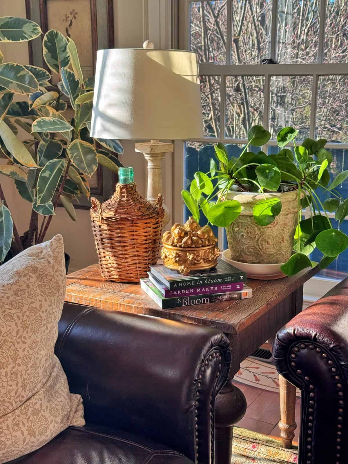 A cozy living room corner with a brown leather armchair, a side table holding a lamp, potted plants, a woven bottle, a golden decorative object, and stacked books, with sunlight streaming through a window.