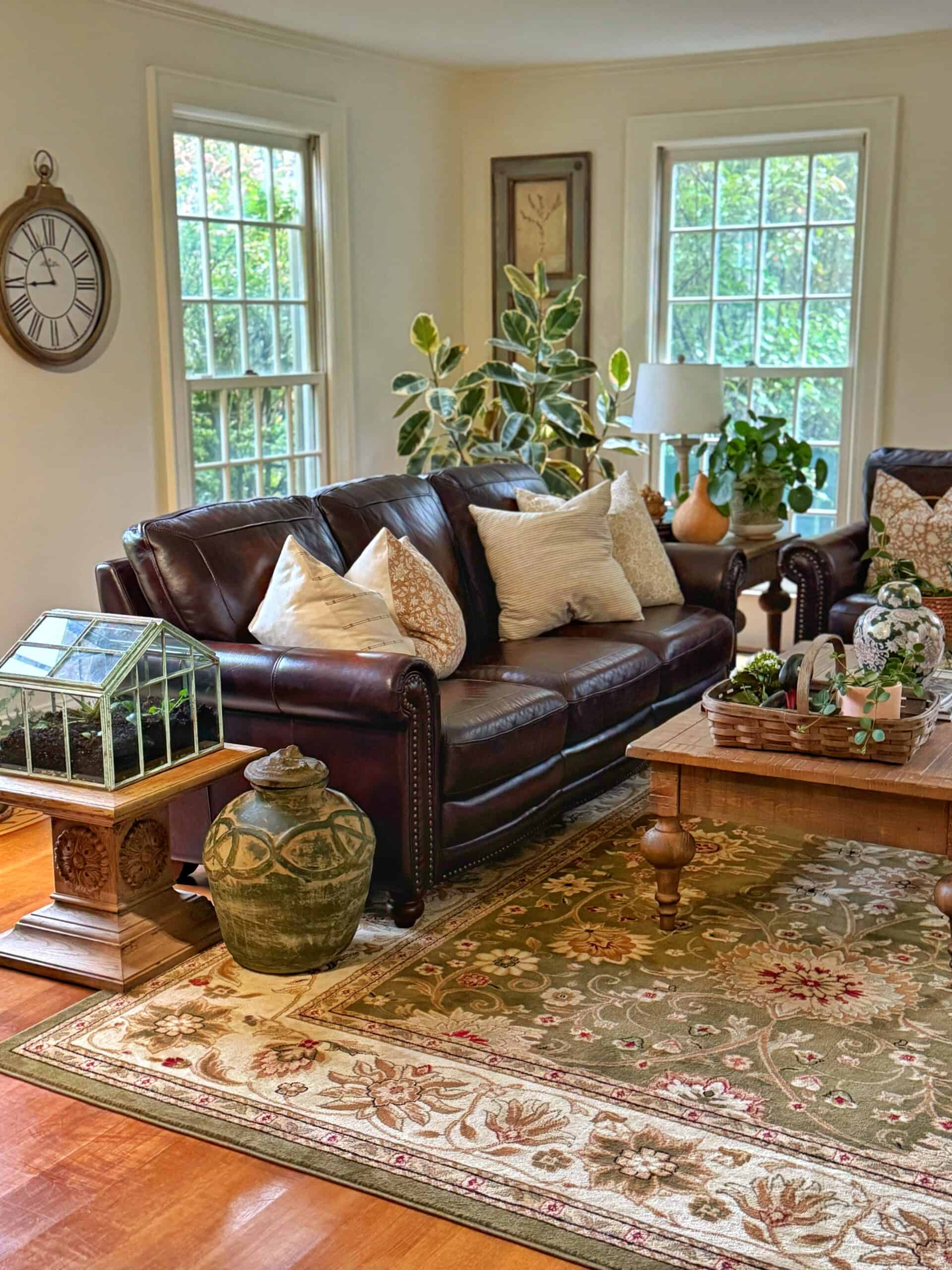 A cozy living room with a brown leather sofa, cream pillows, a wooden coffee table, indoor plants, a decorative clock on the wall, and sunlight streaming through large windows onto a patterned rug.
