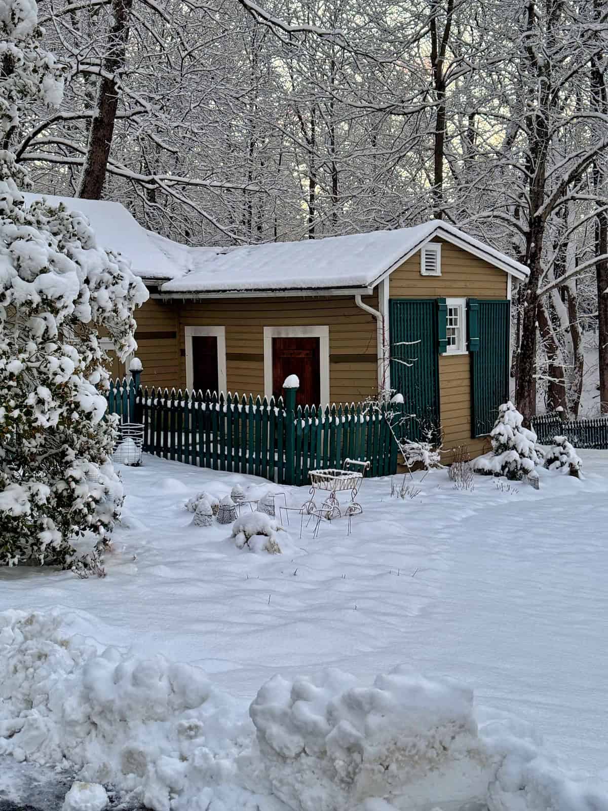 A small tan house with green shutters and a picket fence is surrounded by trees and blanketed in fresh snow. Snow covers the roof, fence, bushes, and ground, creating a peaceful winter scene.