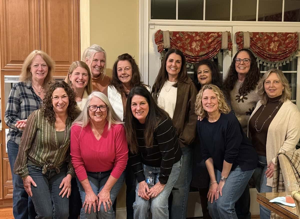 A group of eleven women, smiling and casually dressed, pose together in a warmly lit kitchen in front of a window with patterned curtains. Some are standing, while others are kneeling in front of them.