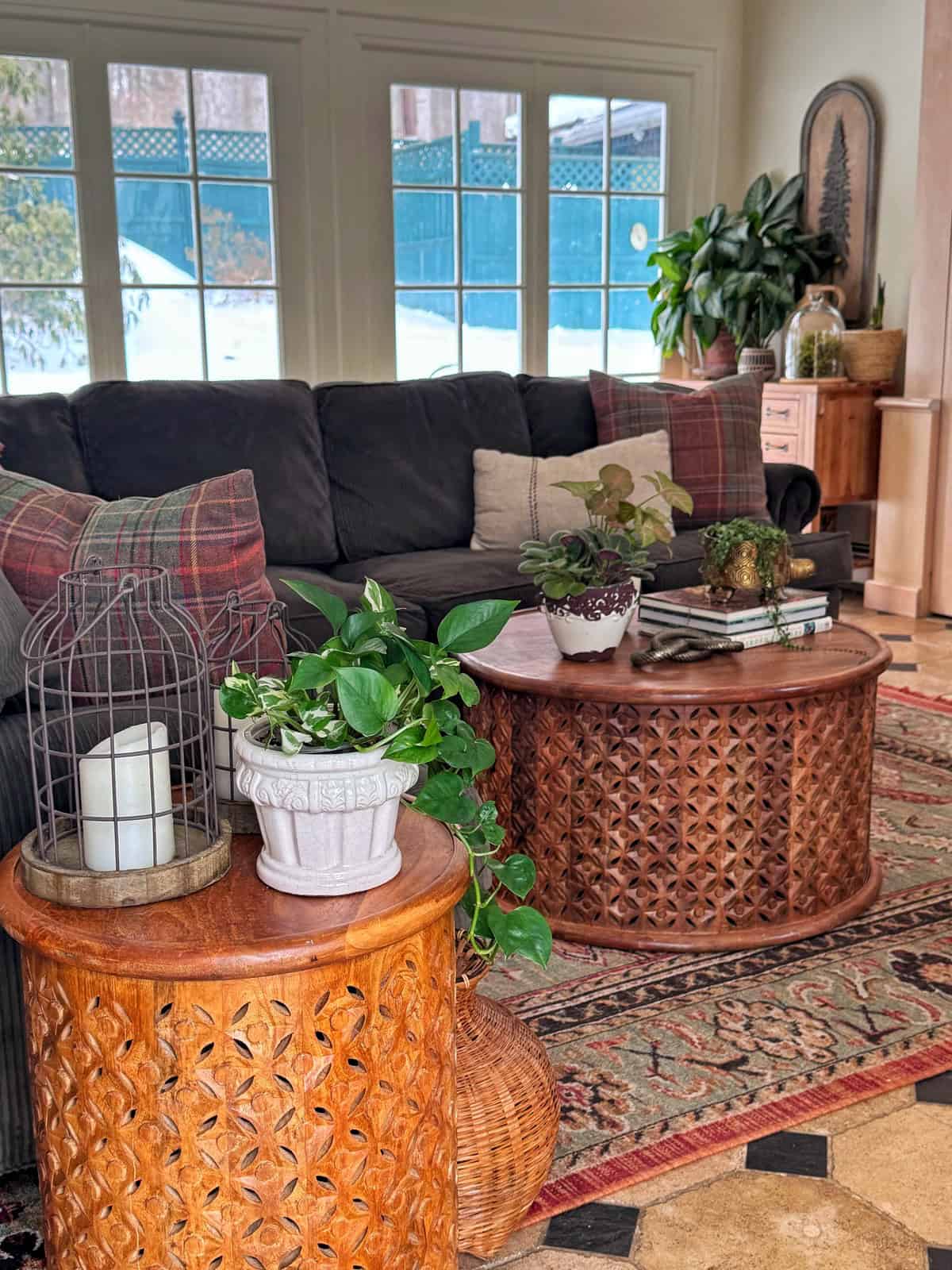 A cozy living room with a dark couch, plaid pillows, and two round wooden tables topped with potted plants, books, and a wire candle holder, next to large windows letting in natural light.