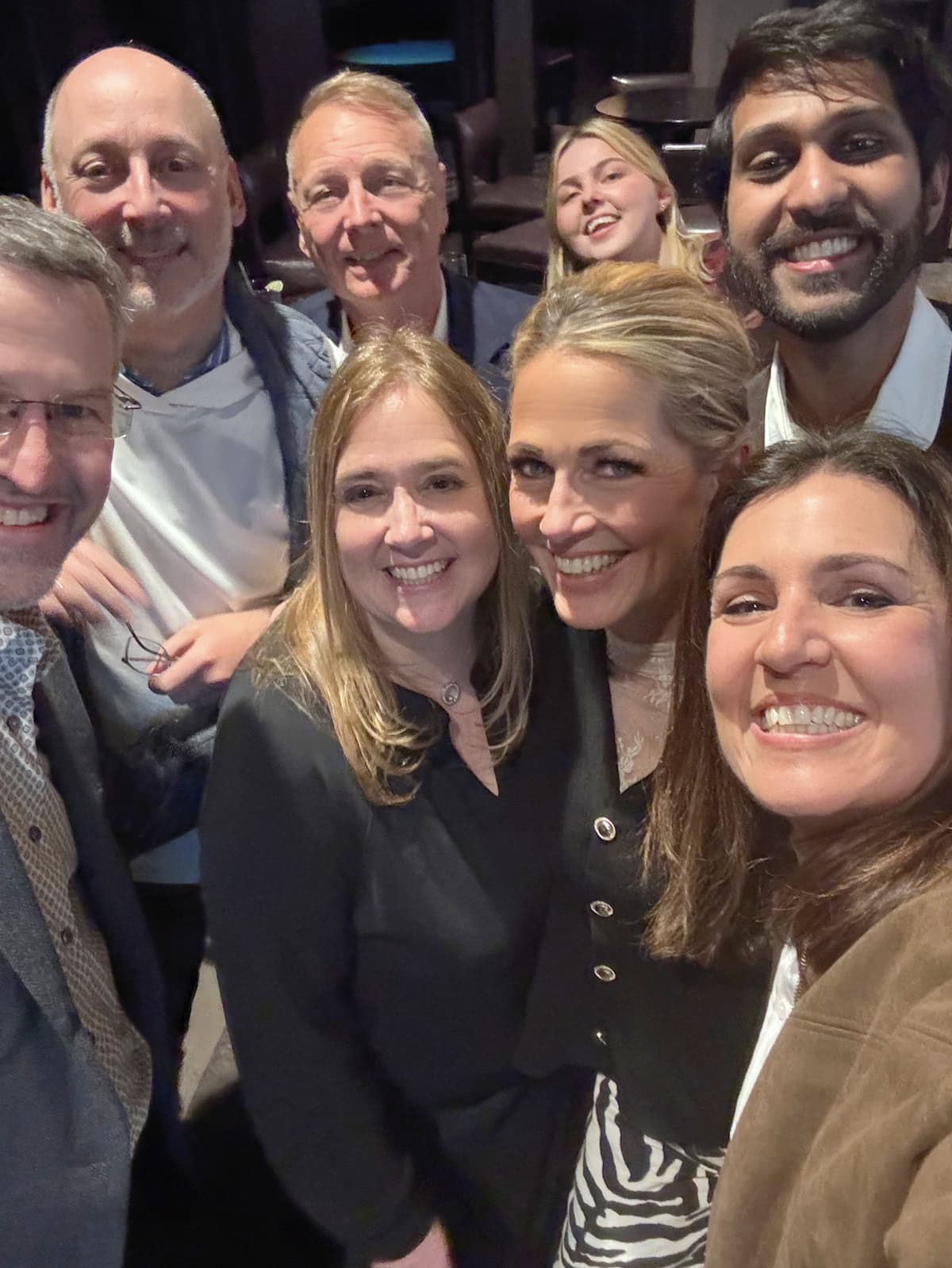 A group of eight adults, both men and women, are standing close together, smiling and taking a selfie indoors. They appear happy and are dressed in smart casual clothing.