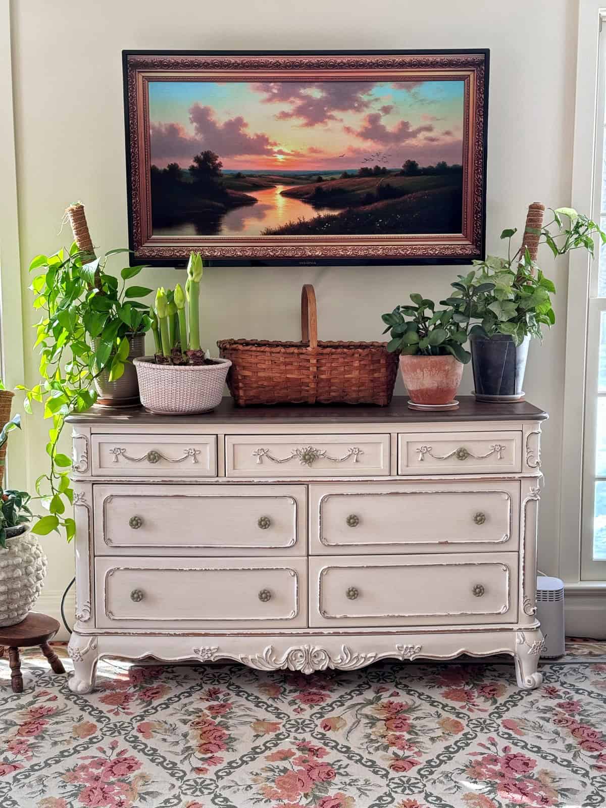 A vintage white dresser is topped with potted plants and a basket. Behind it, a framed sunset landscape painting hangs on a cream wall. Floral-patterned carpet covers the floor. Natural light streams in from the right.