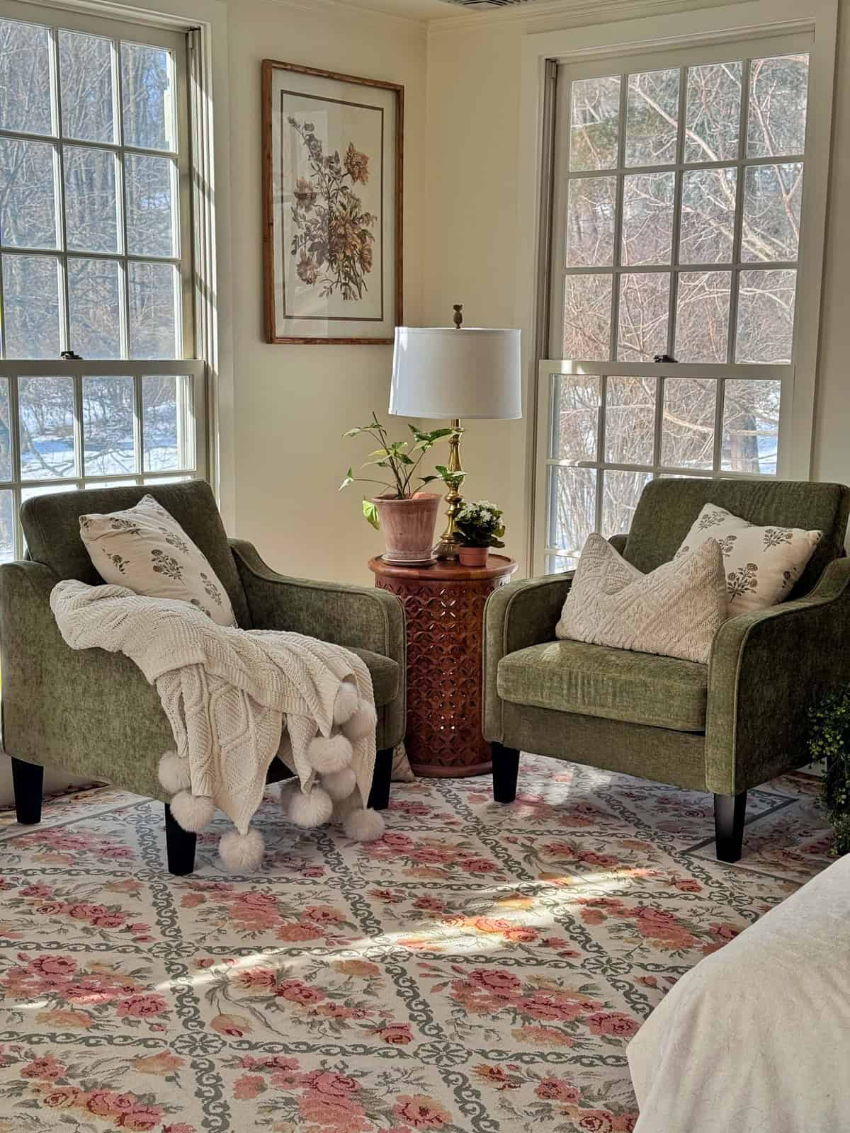 Two green armchairs with patterned pillows sit by large windows in a sunlit room. A knit blanket with pom-poms is draped over one chair. A round side table with a lamp and potted plant sits between the chairs, on a floral rug.