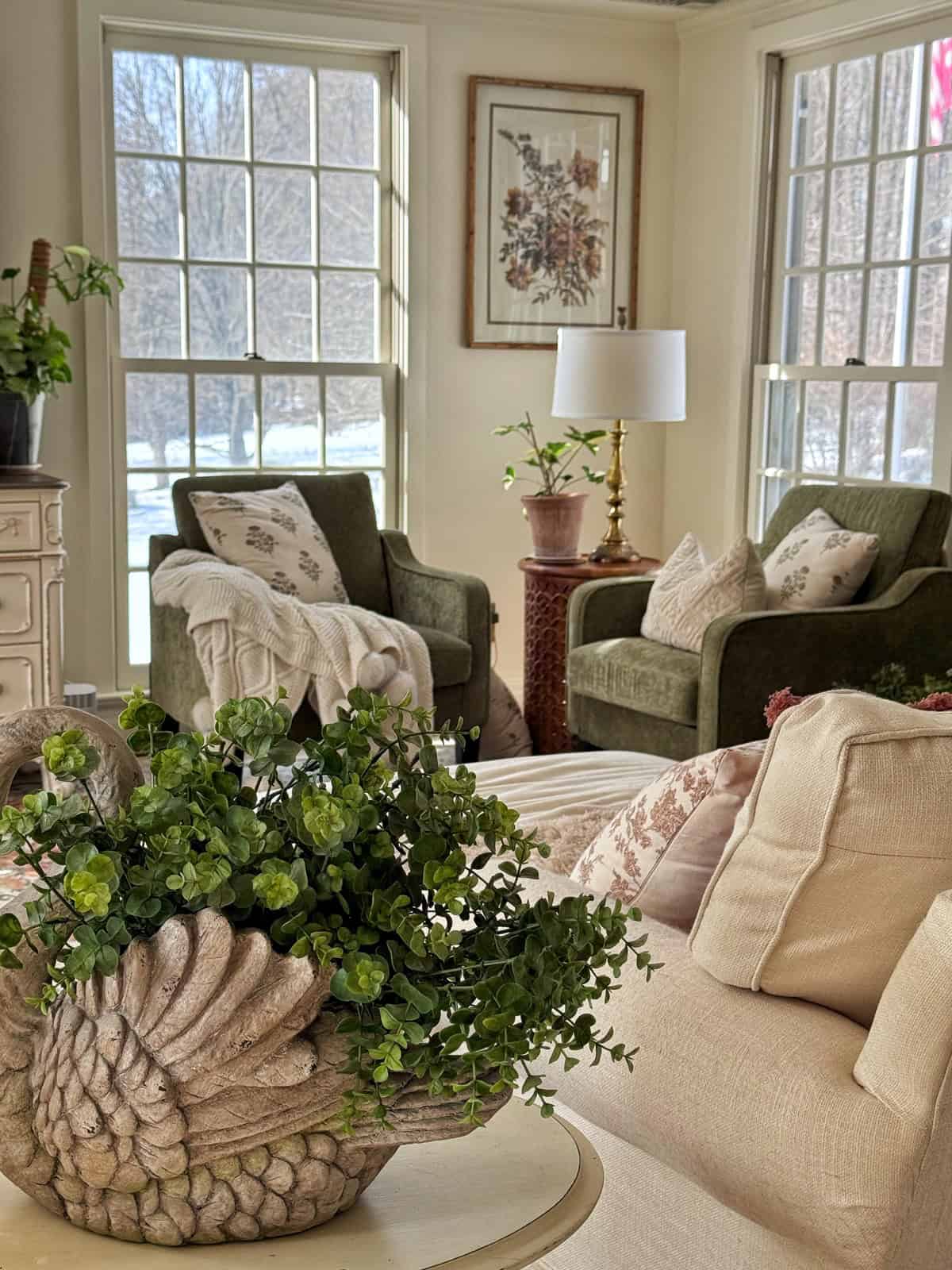 A cozy, sunlit living room with large windows, beige sofas, green armchairs, decorative pillows, houseplants, and a table centerpiece featuring lush greenery in a shell-shaped planter. Snow is visible outside.