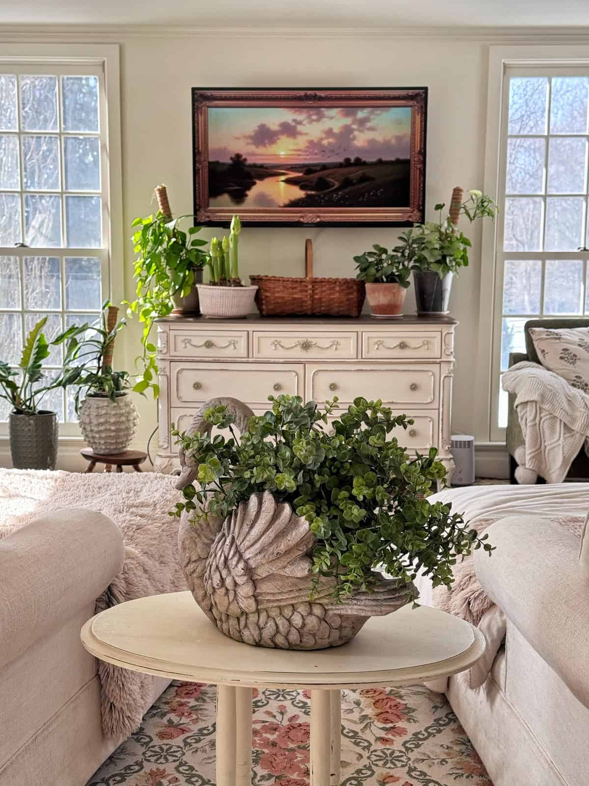 A bright living room with large windows, green plants on a white dresser, a framed sunset photo on the wall, and a swan-shaped planter with greenery on a round table between two sofas.