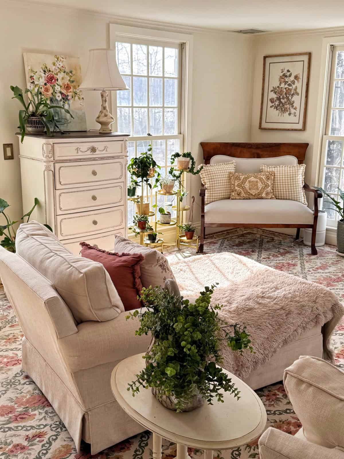 Bright, cozy living room with a beige armchair, ottoman, and loveseat, floral rug, potted plants, a white dresser, and large windows letting in sunlight. Floral artwork and throw pillows add warm, inviting touches.