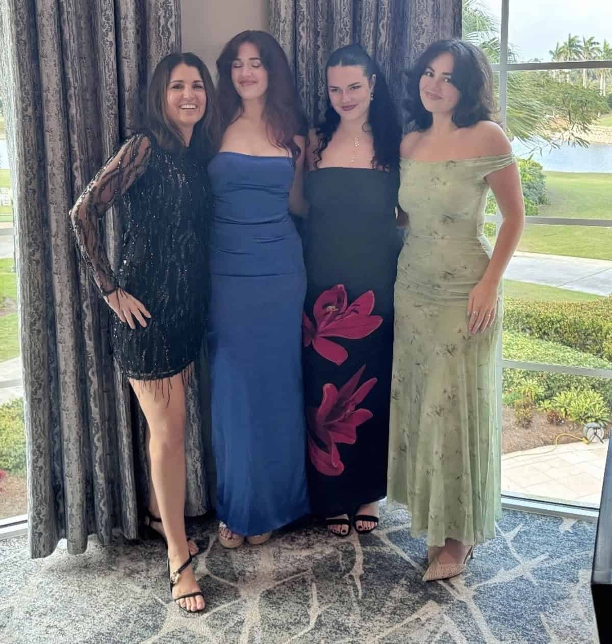 Four women dressed in formal evening gowns stand together, smiling indoors by large windows with a scenic outdoor view of grass, trees, and water in the background.