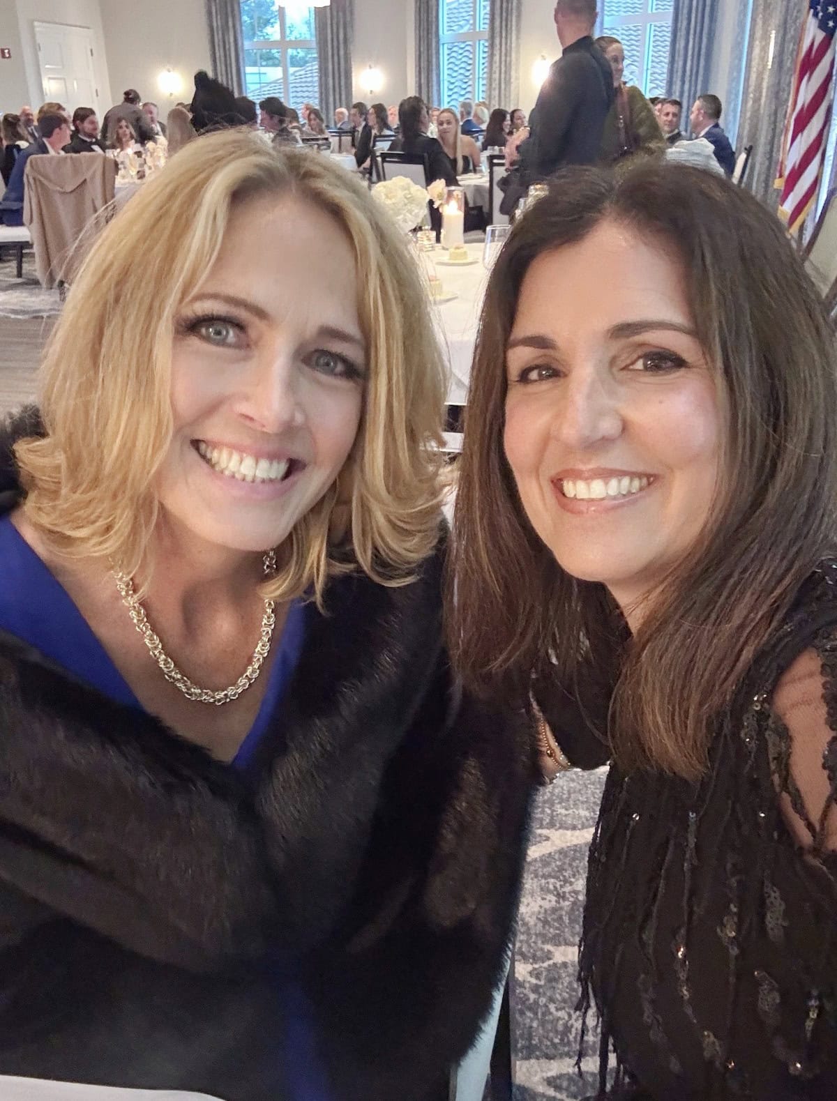 Two women smiling and posing for a selfie at a formal indoor event. Both are dressed elegantly; one wears a black fur stole and pearl necklace, the other a black lace dress. People and tables are visible in the background.