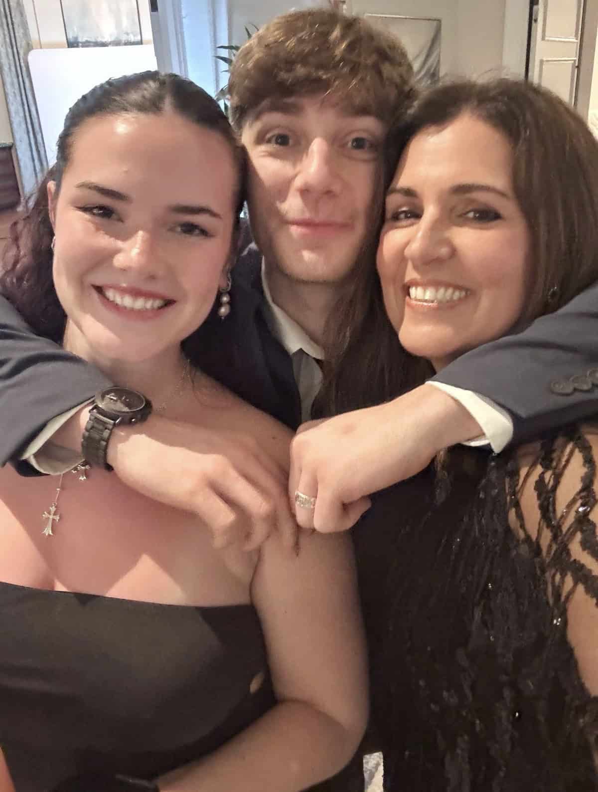 Three people pose closely together, smiling for a selfie at a formal event. The young man in the middle has his arms around the two women, all dressed in elegant attire, looking happy and cheerful indoors.