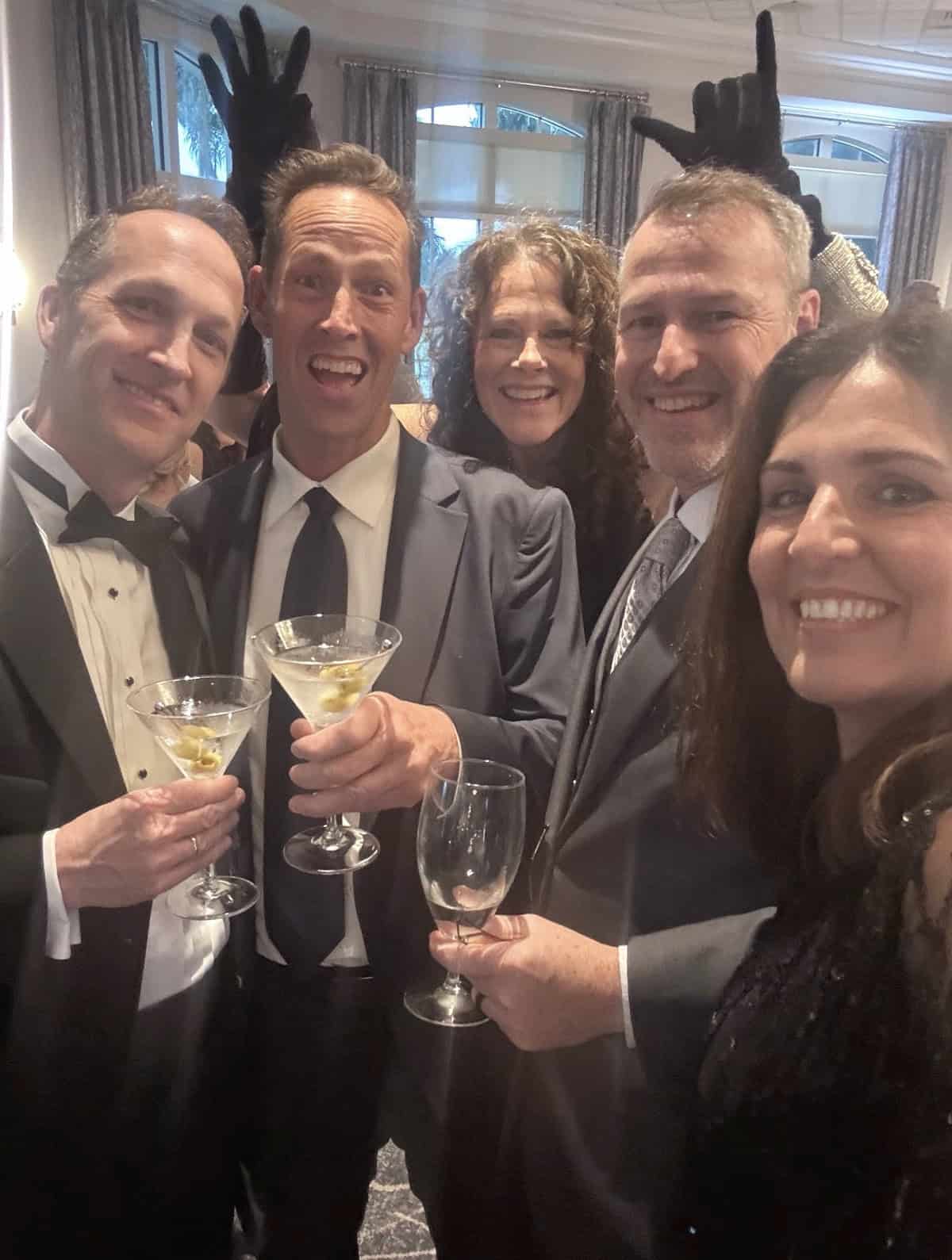Five people dressed formally smile and pose for a selfie at a festive event, holding drinks. Someone in the back playfully makes bunny ears behind another person's head.