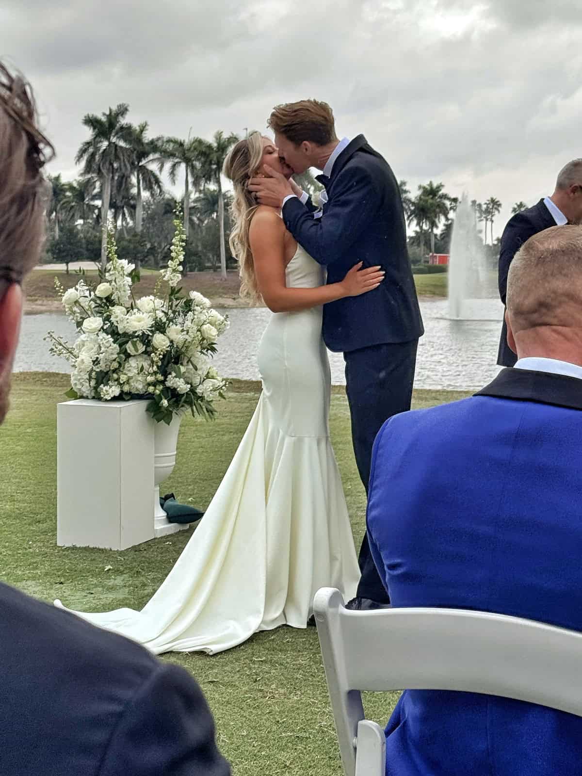 A bride and groom kiss at an outdoor wedding ceremony by a lake, with palm trees and a fountain in the background. The bride wears a white gown, and the groom wears a navy suit. Guests are seated nearby.