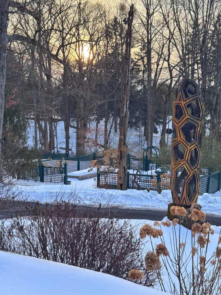 A snowy park scene at sunset with leafless trees, wooden geometric sculptures, a fence, and dried plants in the foreground. The sun is low in the sky, casting a warm glow over the winter landscape.