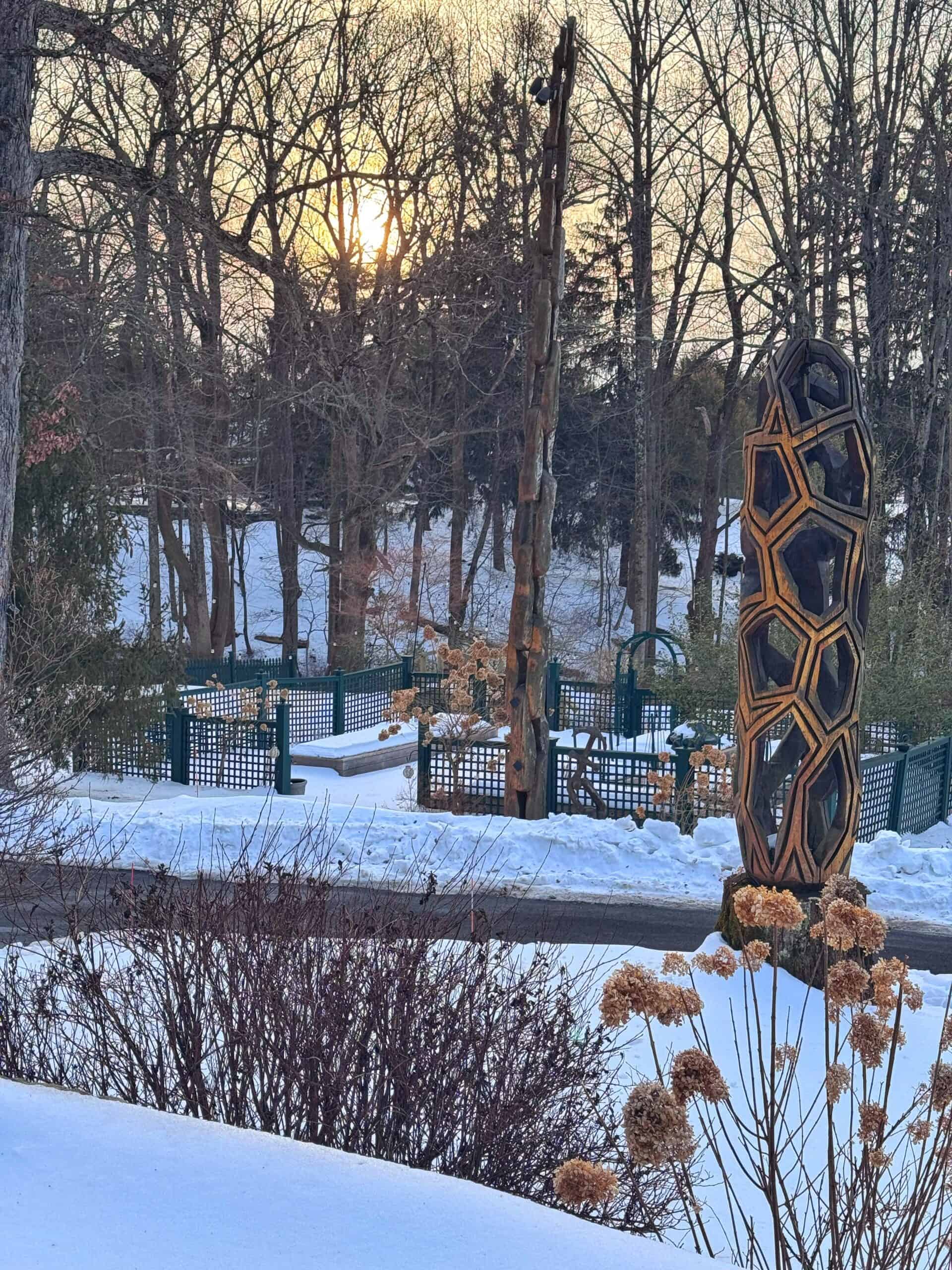 A snowy park scene at sunset with leafless trees, wooden geometric sculptures, a fence, and dried plants in the foreground. The sun is low in the sky, casting a warm glow over the winter landscape.