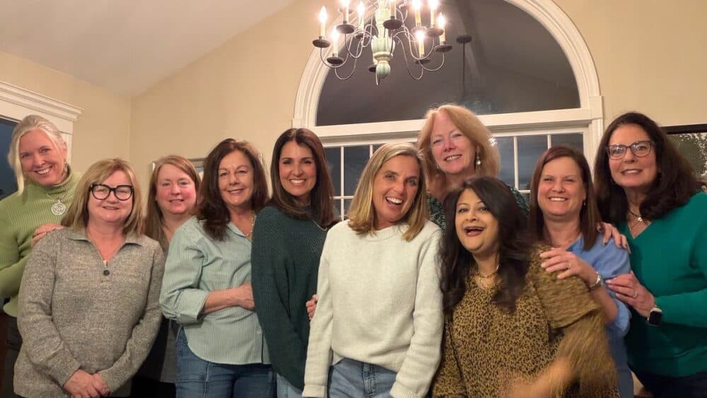 A group of ten women smiling and posing together indoors in front of a window, under a chandelier, with beige walls and casual attire.