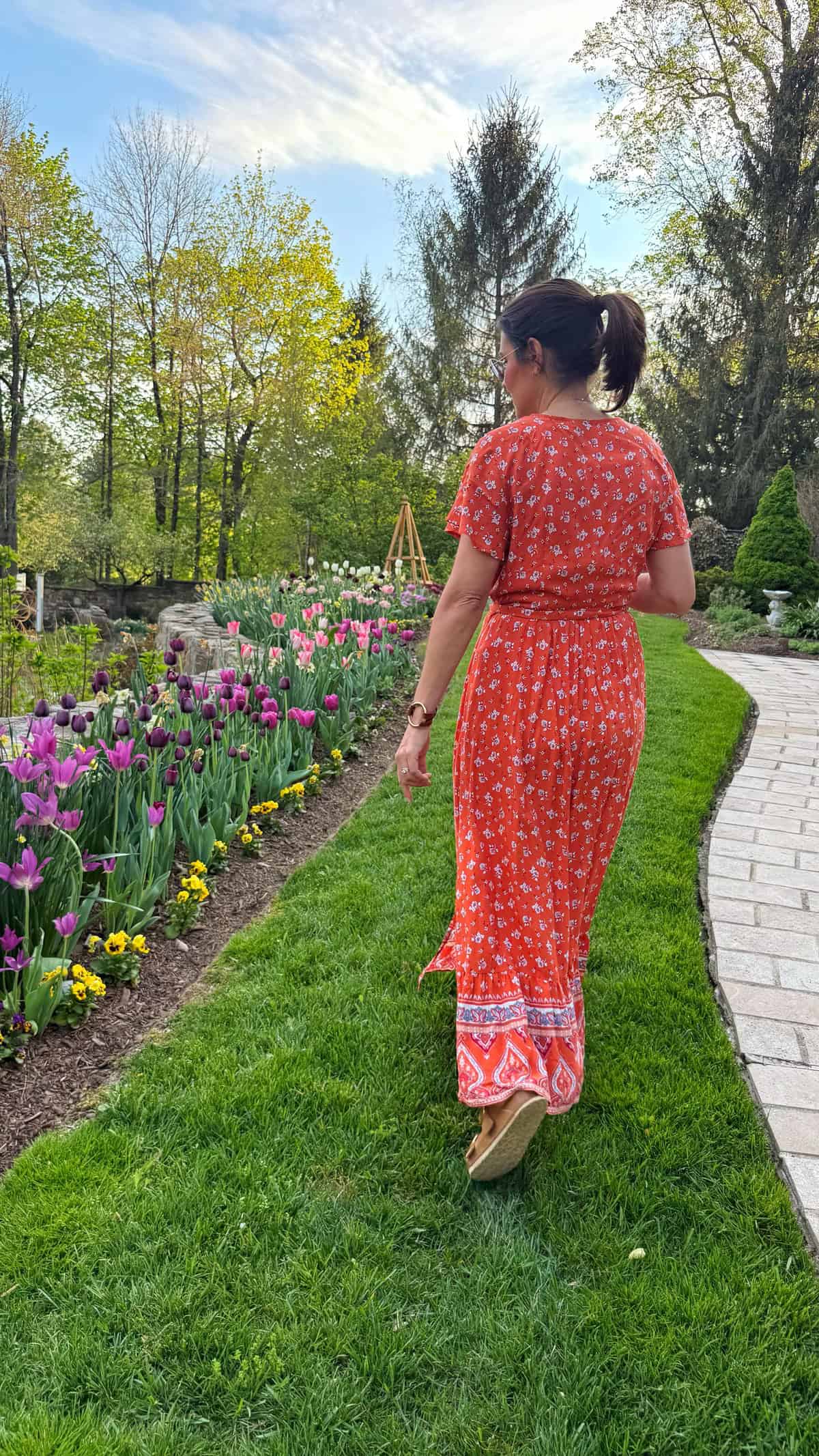 stacy ling in an orange floral boho dress walks along a garden path lined with blooming tulips and yellow flowers on a sunny day, surrounded by green grass and tall trees.