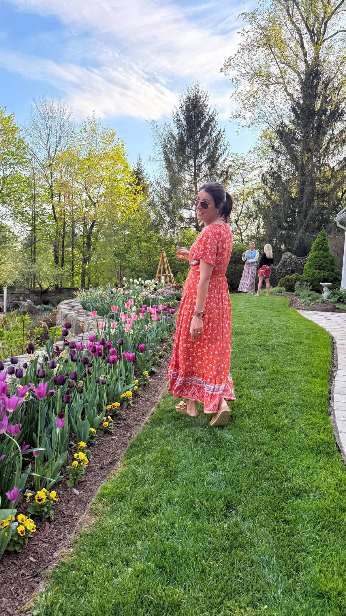 stacy ling in an orange patterned dress and sunglasses stands on a grassy path next to blooming tulips in a garden, with trees and other people visible in the background on a sunny day.