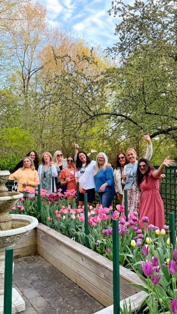 A group of women pose smiling and waving around a raised garden bed filled with colorful tulips in a park, with trees and a fountain in the background under a partly cloudy sky.