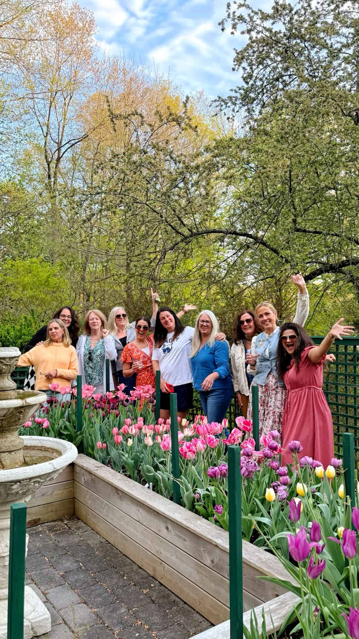 A group of women pose smiling and waving around a raised garden bed filled with colorful tulips in a park, with trees and a fountain in the background under a partly cloudy sky.