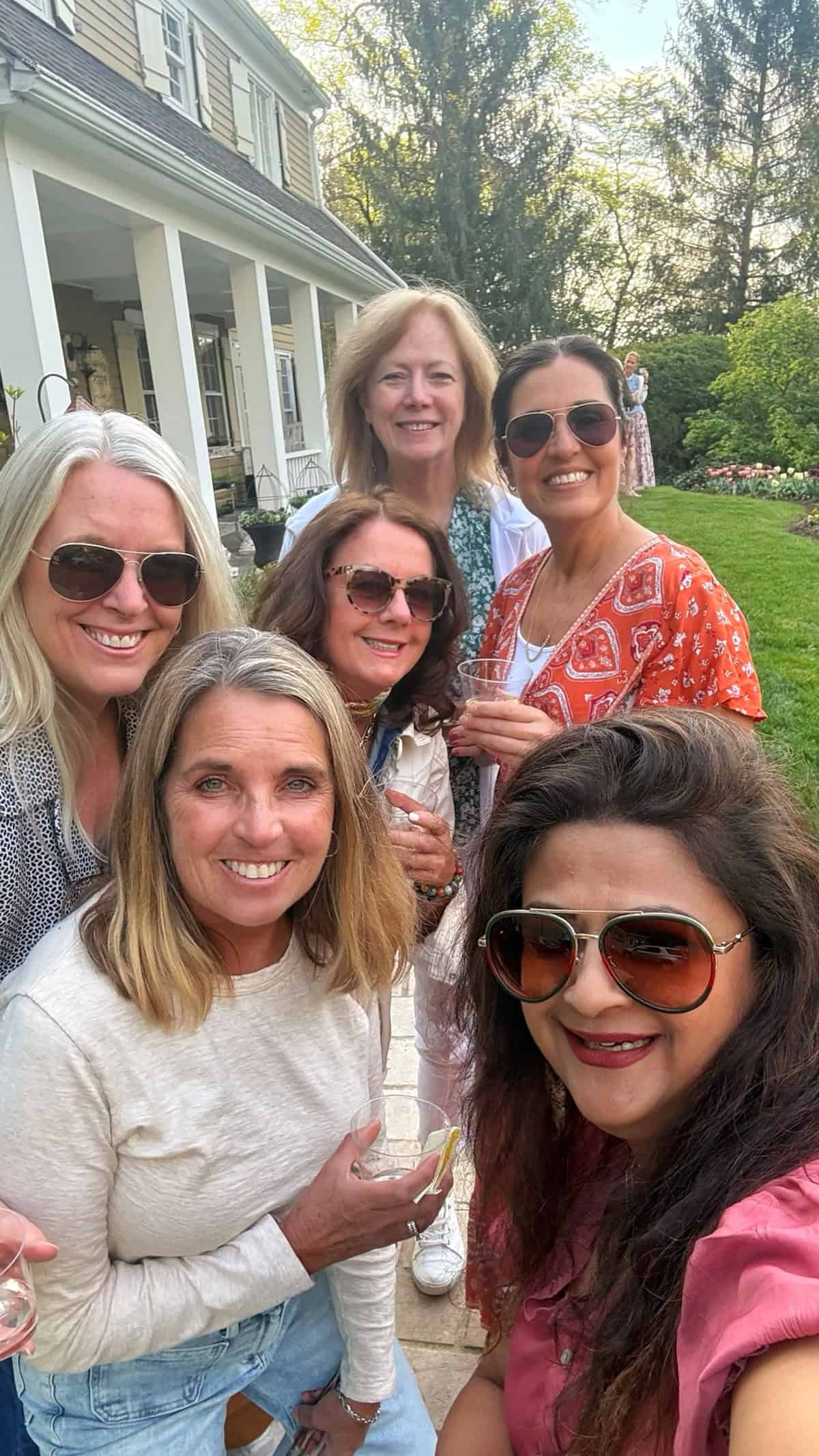 Six women smiling and posing together outdoors in front of a house with a porch and garden. They are wearing sunglasses and casual clothing, enjoying a sunny day.