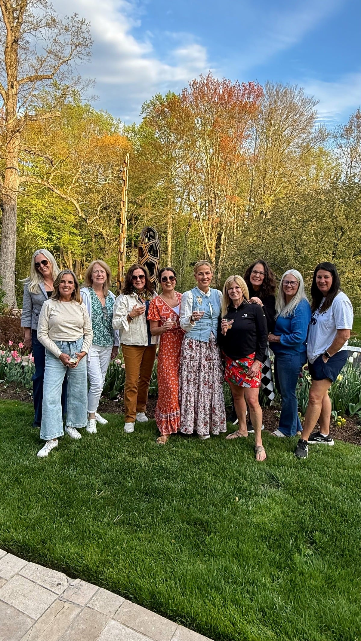 A group of ten women pose together outdoors on a grassy lawn, with trees and flowers in the background under a partly cloudy sky. They are dressed in casual spring clothing and appear to be enjoying a gathering.