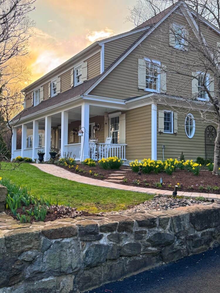 A tan two-story house with white trim and a covered front porch, surrounded by blooming yellow daffodils and a stone retaining wall, at sunset.