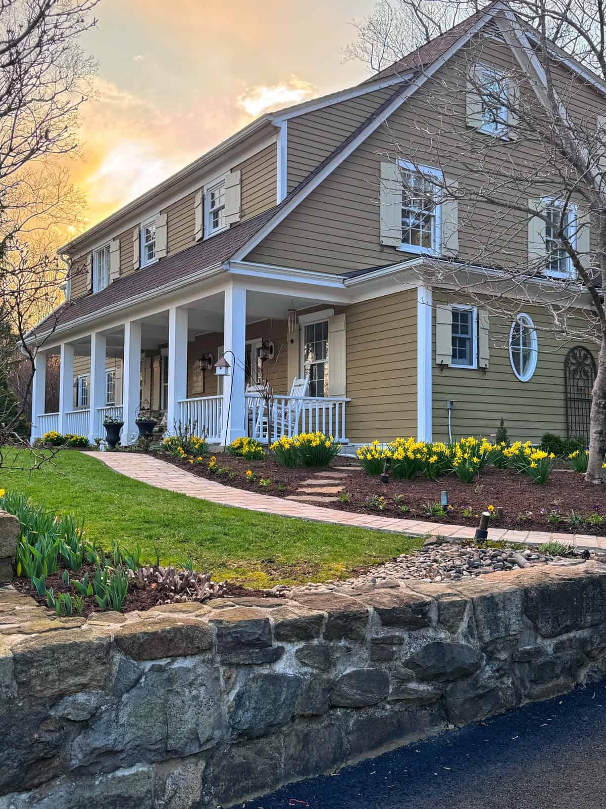 A tan two-story house with white trim and a covered front porch, surrounded by blooming yellow daffodils and a stone retaining wall, at sunset.