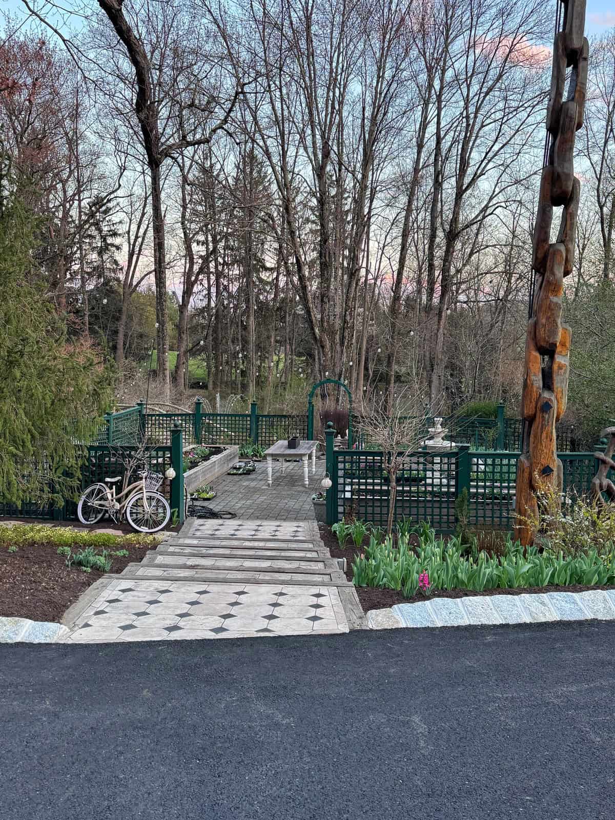 A paved garden path lined with greenery and flowers leads to a bench. A white bicycle is parked to the left, and a tall wooden sculpture stands to the right. Leafless trees fill the background under a dusky sky.