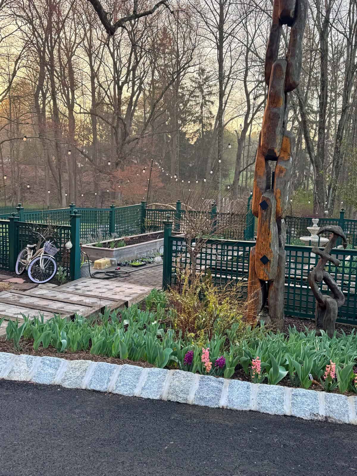 A garden with blooming flowers, a wooden sculpture, string lights, green fencing, a bench, and two bicycles resting on a paved area; tall bare trees fill the background.