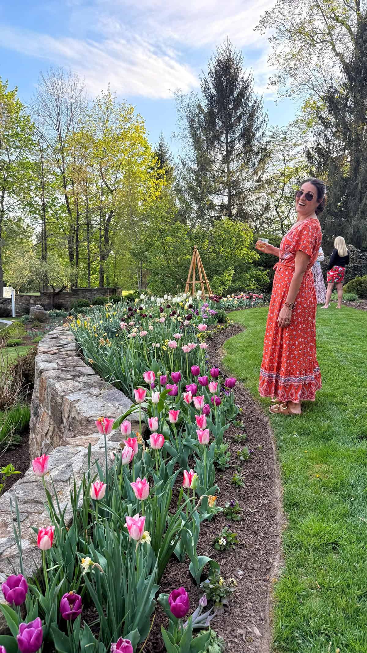 A woman in a red patterned dress stands smiling beside a garden bed filled with pink and purple tulips. She wears sunglasses and sandals. Trees and another person appear in the background on a sunny day.