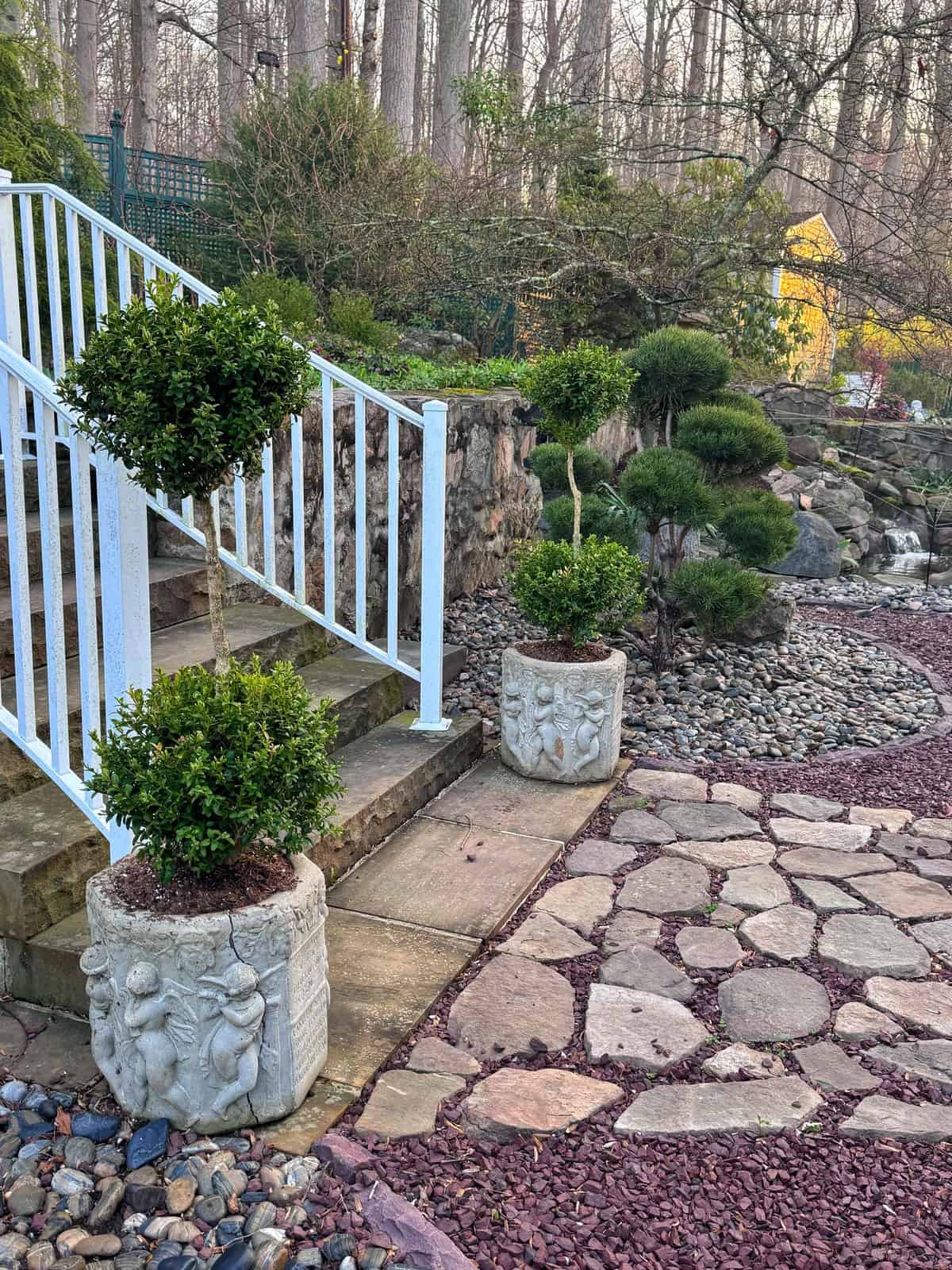 Two potted topiary bushes sit beside stone steps with a white railing. A stone path leads through a landscaped zen garden with rocks, shrubs, and trees. A yellow structure is visible in the background.